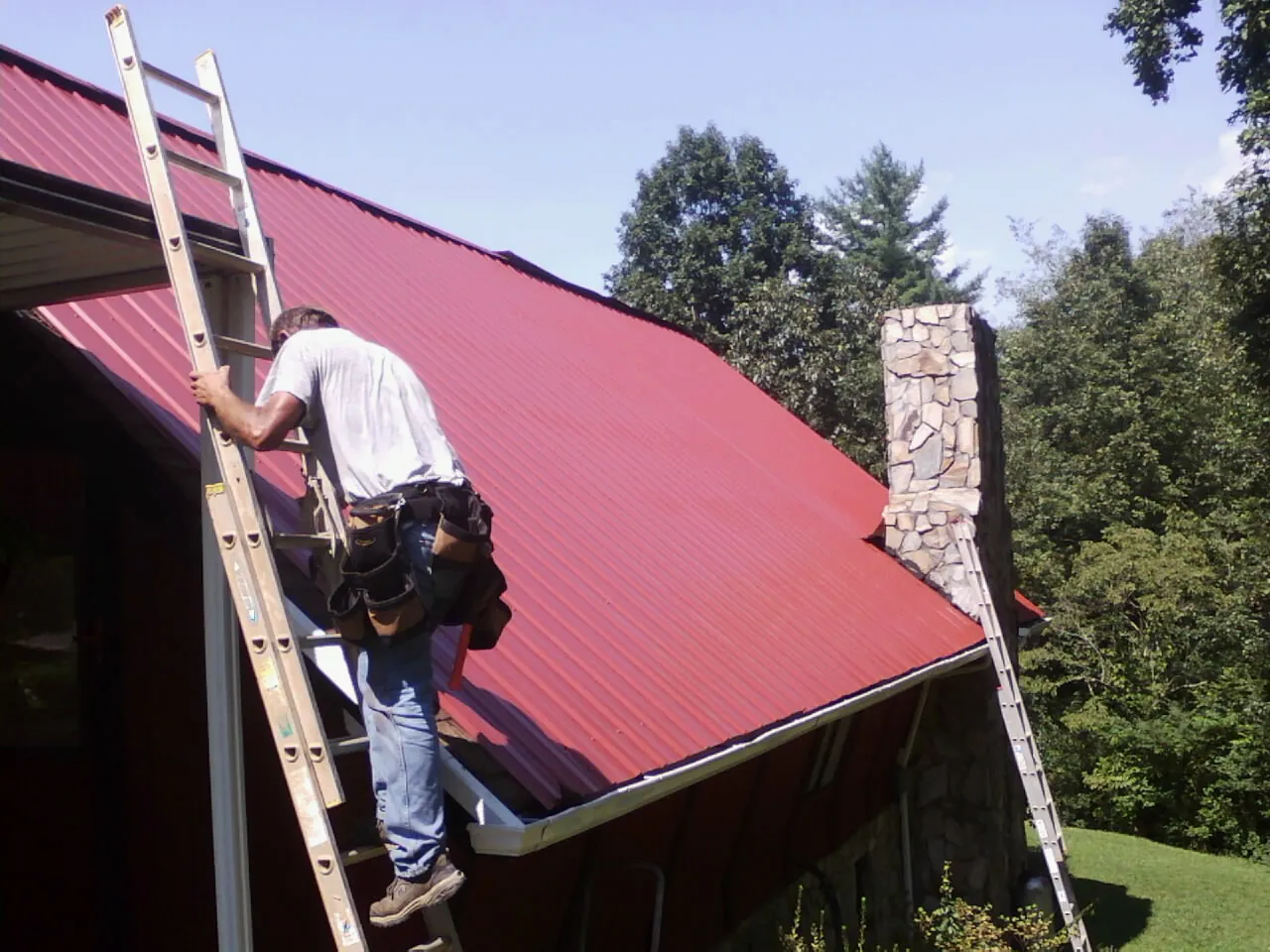 Roofer on a ladder beside a red metal roof and stone chimney — Soffit & Fascia Repair by Roofing Brothers in Prophetstown